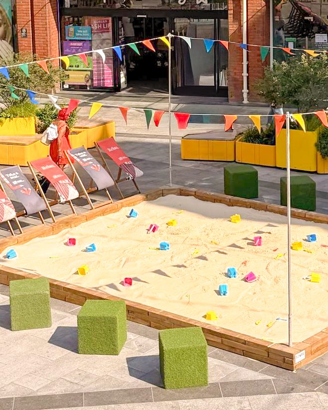 Timber framed sandpit with branded deckchairs and grass seating cubes in a shopping centre square